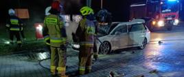 Strażacy stoją przy ugaszonym samochodzie osobowym koloru białego. W tle po prawej stronie stoi samochód pożarniczy