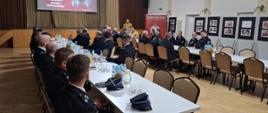 Strażacy ubrani w granatowe mundury galowe, oraz pisakowe mundury siedzą przy stolikach, w tyle rolap z logiem Państwowej Straży Pożarnej na tylnej ścianie kotary oraz ekran z wyświetloną prezentacją