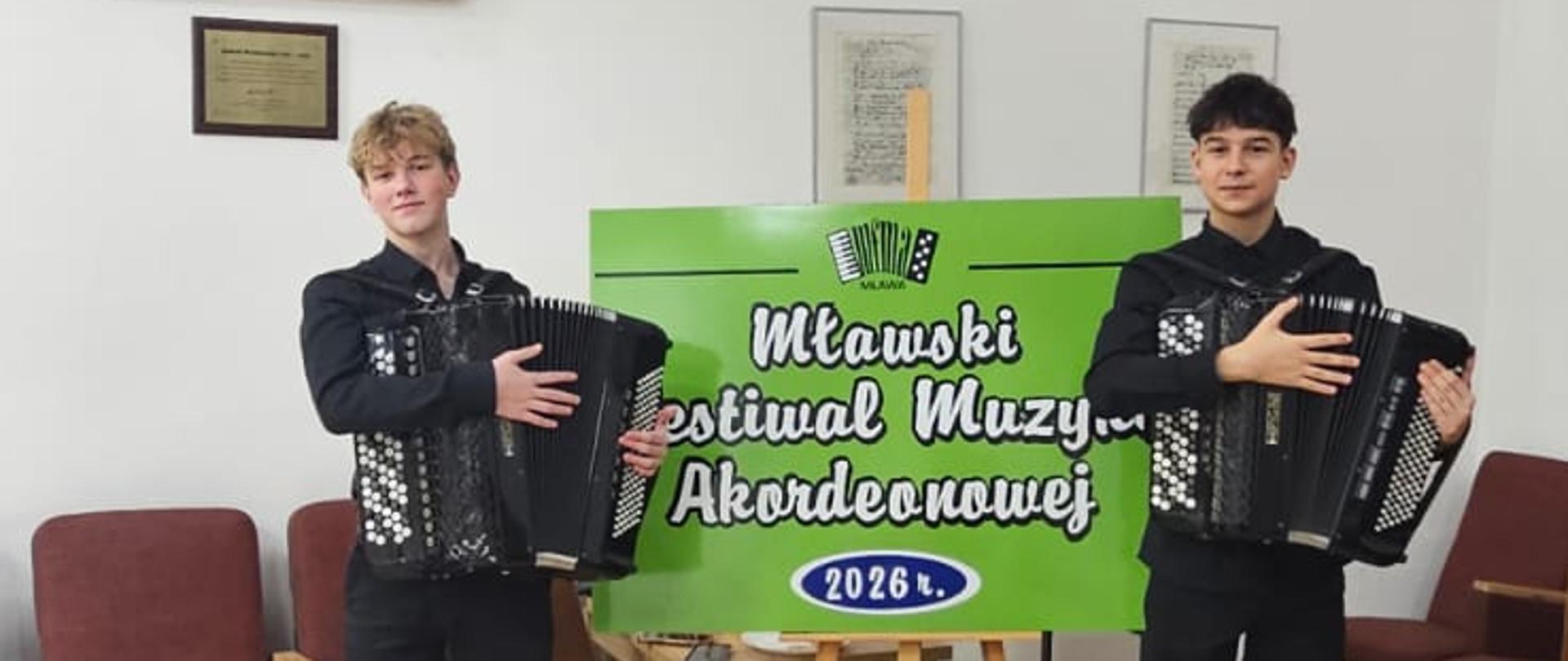 Dwóch uczniów ubranych na czarno stoi na drewnianej podłodze, w tle dekoracje, trzymają akordeony.