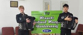 Dwóch uczniów ubranych na czarno stoi na drewnianej podłodze, w tle dekoracje, trzymają akordeony.