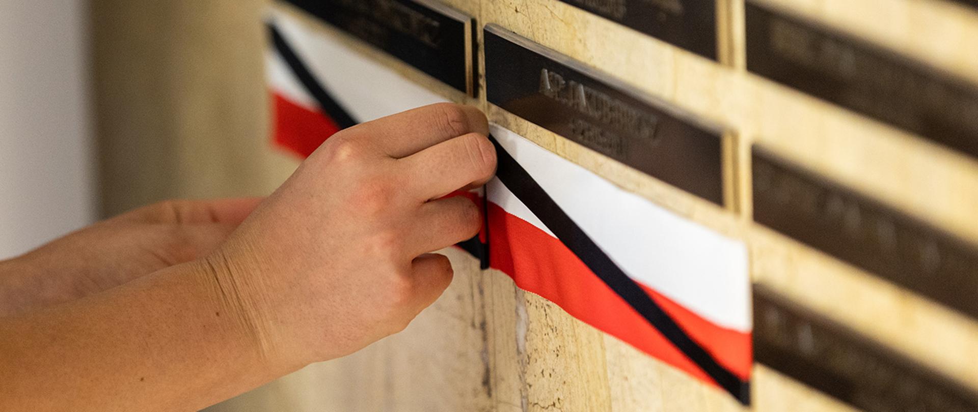 Zdjęcie przedstawiające dłonie, które odsłaniają tabliczkę epitafijną. 