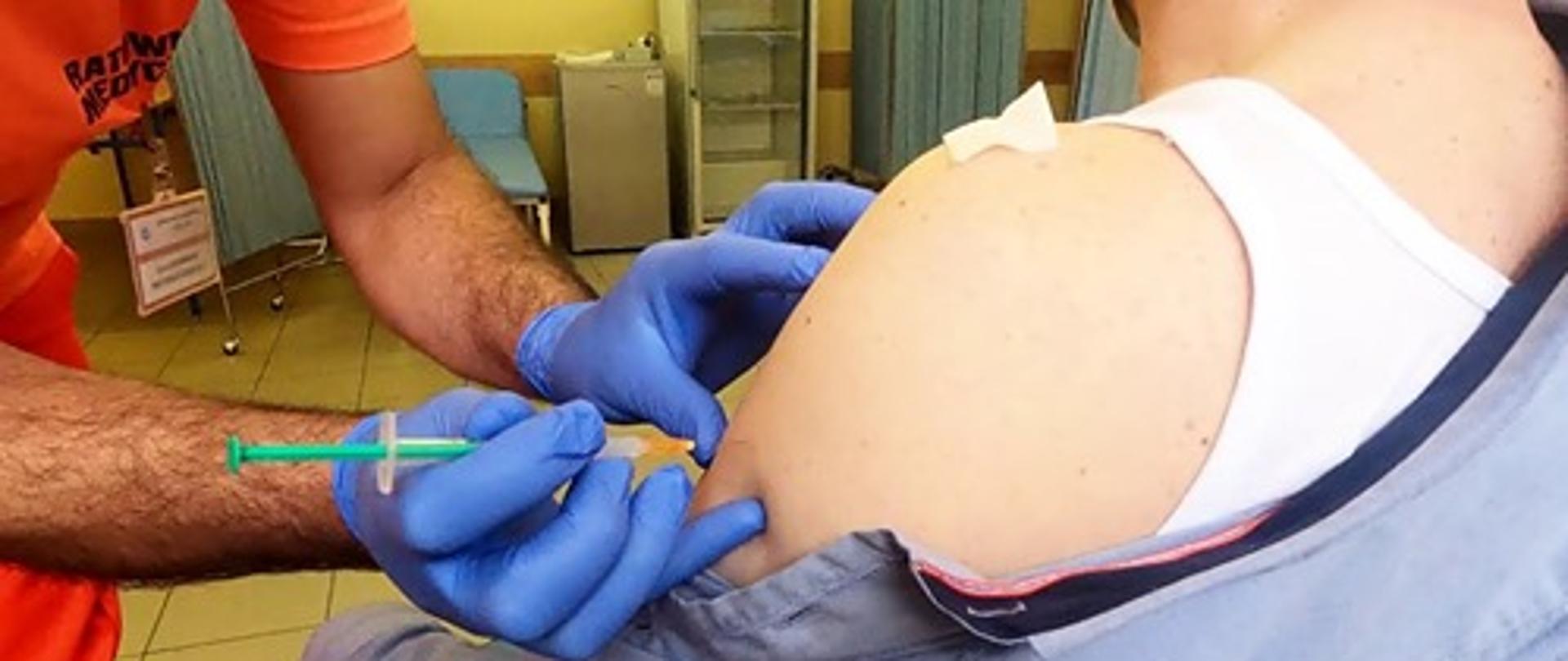 Na pierwszym planie ratownik medyczny w czerwonym podkoszulku i w niebieskich rękawiczkach szczepi pacjenta w lewe ramię. Szczepiony siedzi tyłem, ubrany w biały podkoszulek i niebieską koszulę zsuniętą z ramienia. Na drugim planie widać szklaną szafkę, lodówkę, kozetkę i dwa parawany.