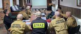Sala wykładowa. Wokół stołu siedzi osiem osób – strażacy w umundurowaniu służbowym i koszarowym – wszyscy plecami do fotografa ze wzrokiem skierowanym na ekran, na którym wyświetlany jest slajd. 