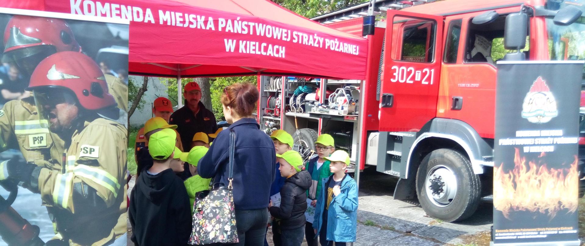 Zdjęcie przedstawia stoisko Państwowej Straży Pożarnej, przy którym skupiła się grupa dzieci wraz z opiekunami. Strażacy edukują najmłodszych a po prawej stronie stoi wóz strażacki.
