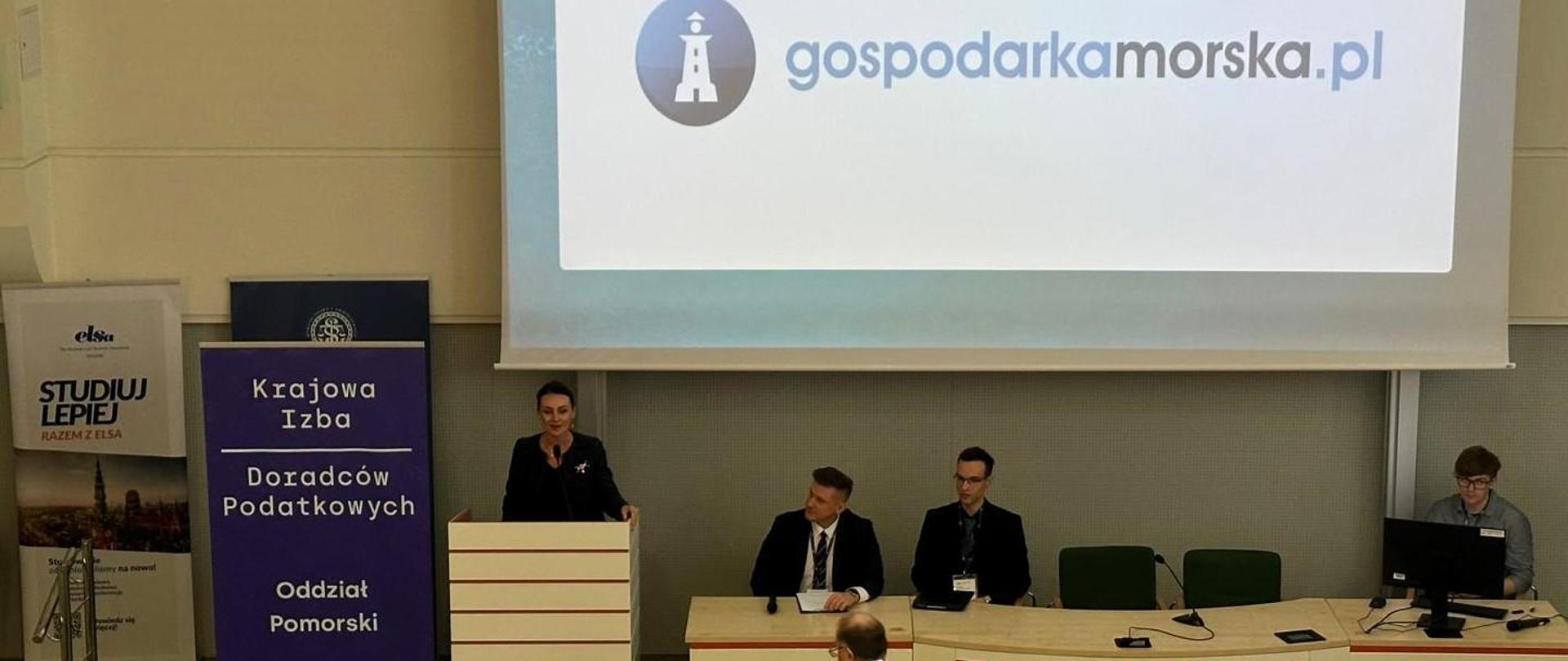 duża sala, kilka osób siedzi za dużym stołem, jedna osoba przemawia, za ni,mi napis gospodarka morska