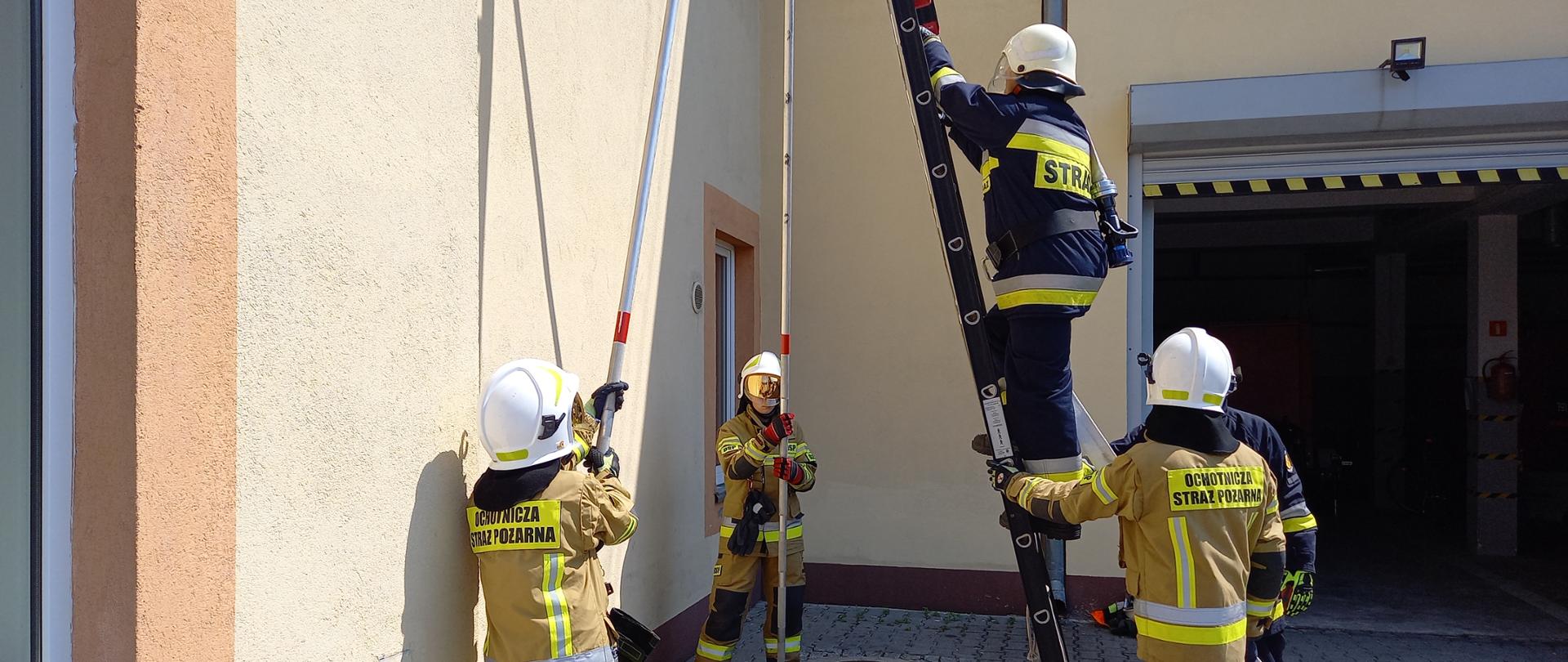 Pięcioro strażaków OSP podczas wykonywania zadania związanego z wprowadzeniem linii wężowej na strażacką drabinę. Dwóch strażaków zabezpiecza drabinę trzymając podpory, kolejnych dwóch zabezpiecza drabinę i jeden strażak wchodzi na drabinę z linią wężową.