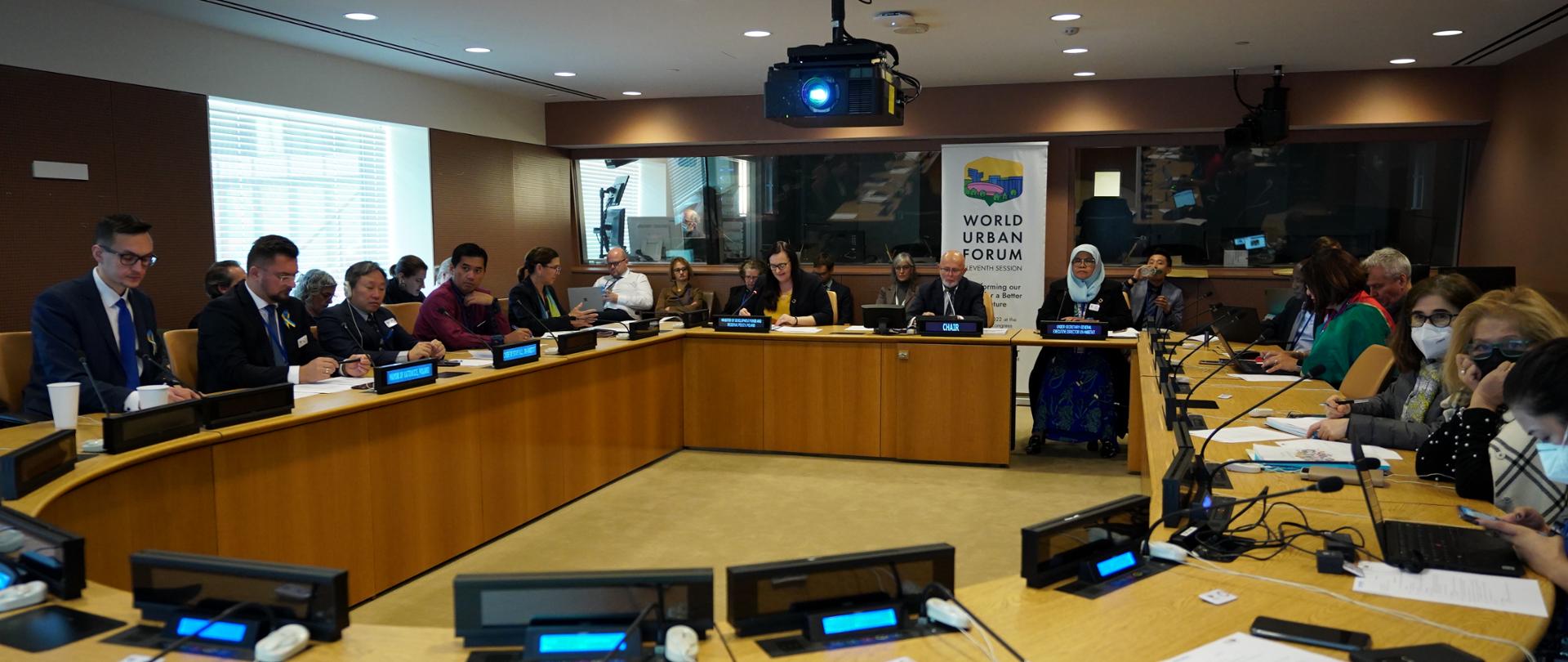 Grupa osób siedzi przy stole z mikrofonami i wyświetlaczami