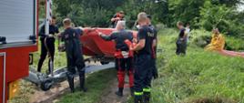 Fotografia przedstawia dziesięć osób na terenie zbiornika wodnego na rzece Bzura. Pięciu ratowników odczepia łódkę ratowniczą barwy czerwonej od przyczepki, za nimi stoi i obserwuje sytuację kobieta, jeden ratownik trzyma w rękach elementy wyposażenia łodzi i patrzy na pięciu ratowników przed nim, ubrany w koszulkę z krótkim rękawkiem z napisem STRAŻ na plecach i w ciemnogranatowe spodnie na szelki z odblaskami na nogawkach, bez nakrycia głowy, buty z wysoką cholewką, dwóch ratowników stojących przy łodzi ubrani są w buty wodery, dwóch ma pianki do nurkowania, jeden czarną drugi czerwoną, wszyscy stoją bez nakrycia głowy, po prawej stronie zdjęcia jeden ratownik ubrany w buty wodery i koszulkę na krótki rękaw obserwuje sytuację dookoła, za nim na pontonie barwy czerwonej siedzi ratownik z grupy poszukiwawczej ratownictwa wodnego w kombinezonie barwy pomarańczowej, w oddali stoi kobieta. W lewej części zdjęcia na pierwszym planie widać fragment pojazdu straży pożarnej barwy czerwonej do którego przypięta jest przyczepka łódki. Dookoła drzewa, trawa, teren lekko podmokły, świeci słońce.