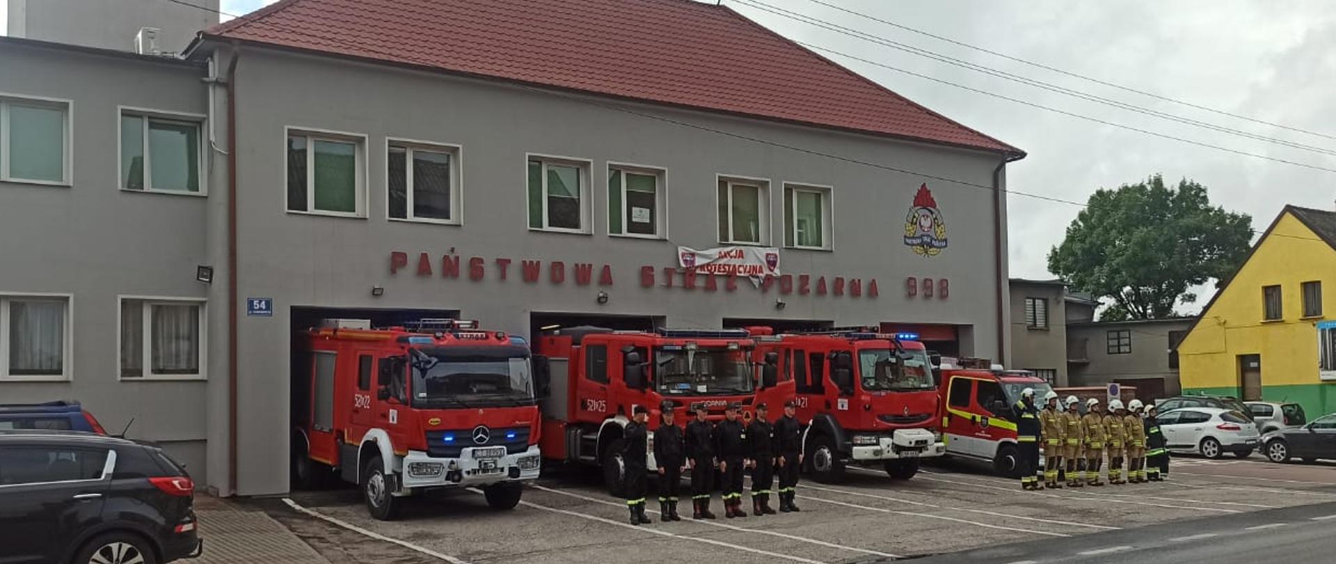Zdjęcie wykonano w ciągu dnia przed budynkiem Komendy Powiatowej Państwowej Straży Pożarnej w Sępólnie Krajeńskim. Strażacy Państwowej i Ochotniczej Straży Pożarnej ubrani w ubrania specjalne i hełmy oraz w ubrania koszarowe stoją na baczność w szeregu przed samochodami ratowniczo- gaśniczymi stojącymi przed garażami. 