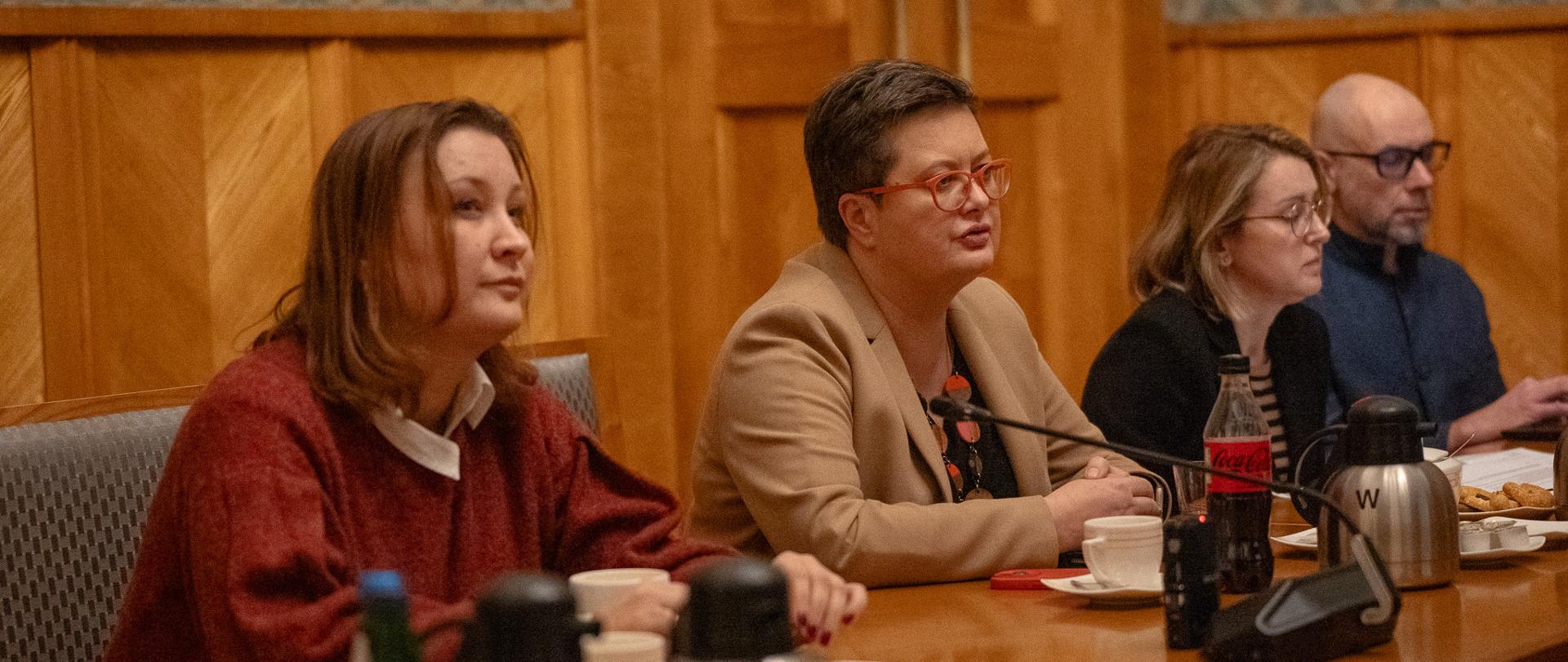 Trzy kobiety i mężczyzna siedzą przy stole. Przed sobą mają mikrofony. Jedna z kobiet mówi do mikrofonu.