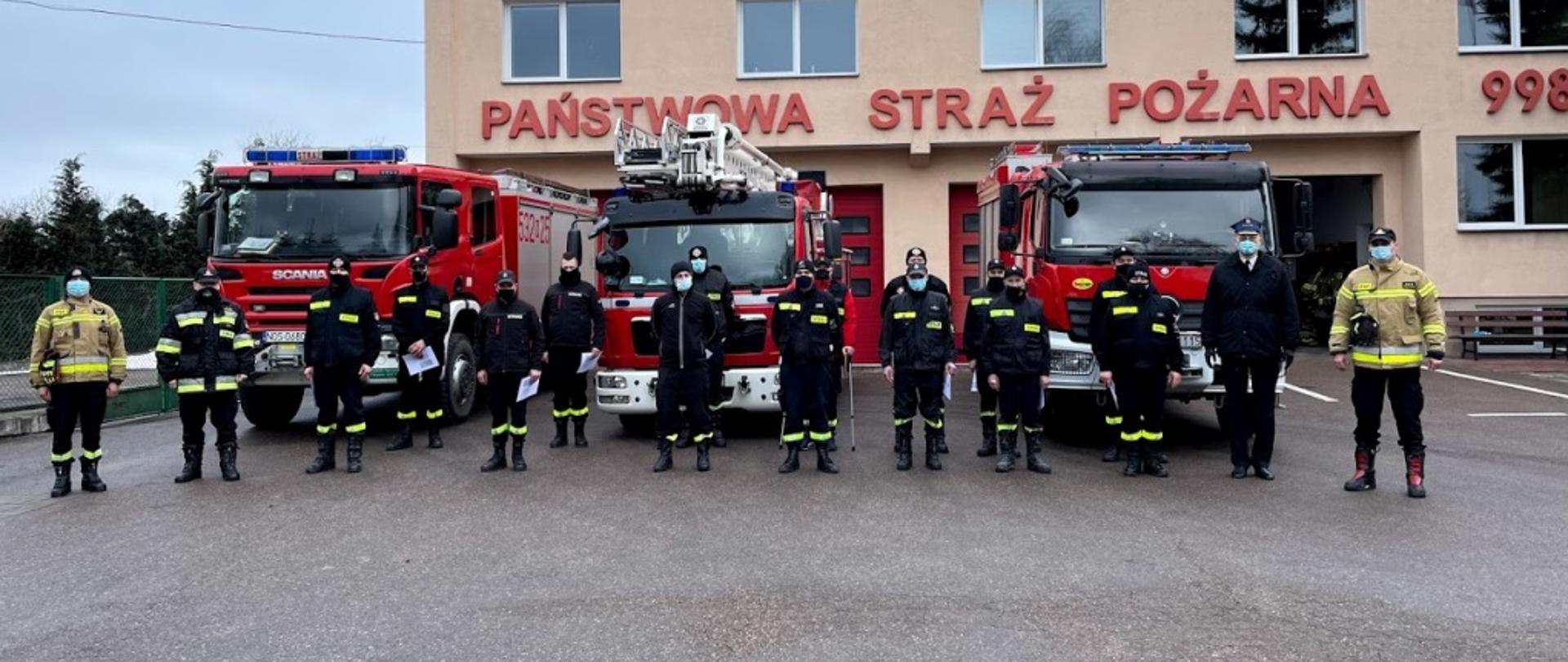 Druhowie ustawieni w dwuszeregu na tle pojazdów pożarniczych i Jednostki Ratowniczo-Gaśniczej w Morągu.