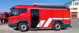 Widok na prawy bok nowoczesnego pojazdu strażackiego Rosenbauer, kolor czerwony, samochód stoi na betonowym placu Szkoły Podoficerskiej.
