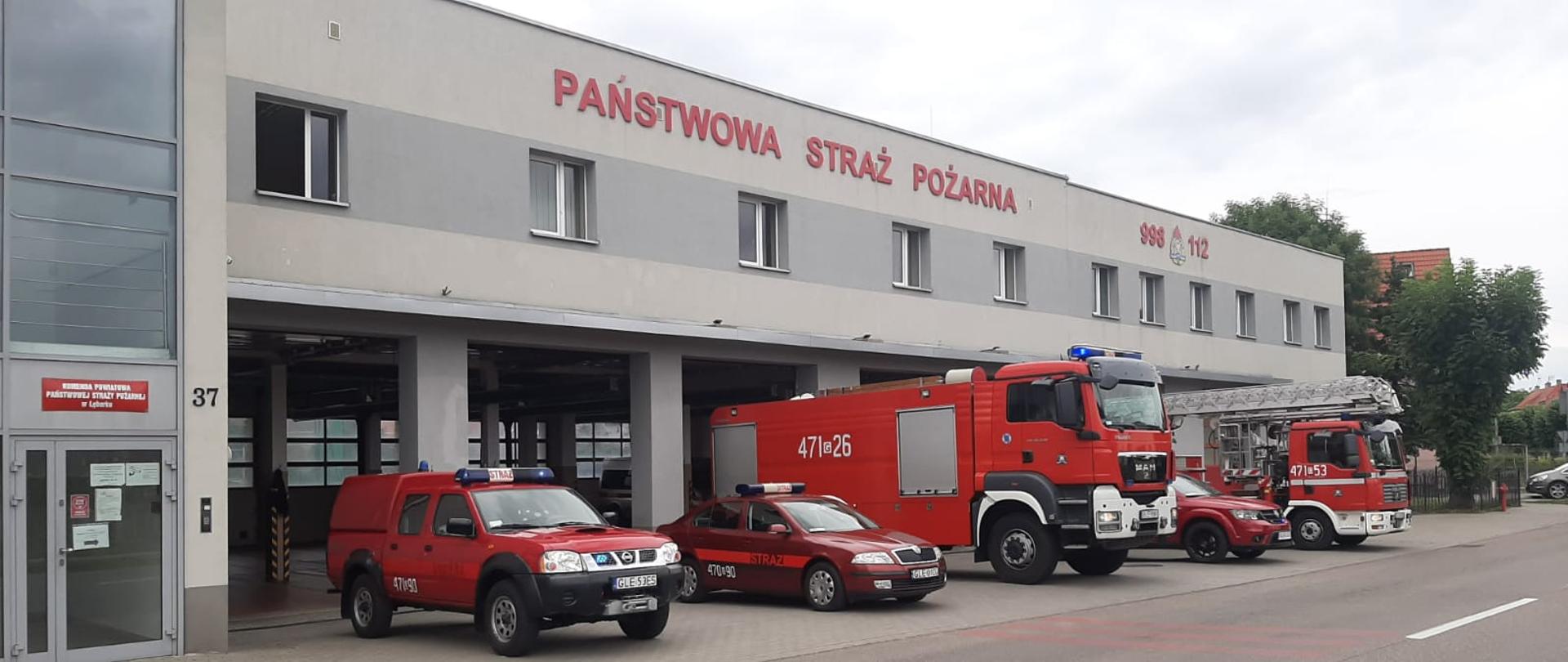 Pięć samochodów Komendy Powiatowej Państwowej Straży Pożarnej w Lęborku z włączonymi sygnałami świetlno-dźwiękowymi wystawione przed otwartymi bramami garażowymi, by przez jedną minutę upamiętnić wydarzenia i Bohaterów biorących udział w walce o Niepodległą Polskę - 77. rocznicę wybuchu Powstania Warszawskiego. Z lewej strony budynku widoczne wejście do komendy. 