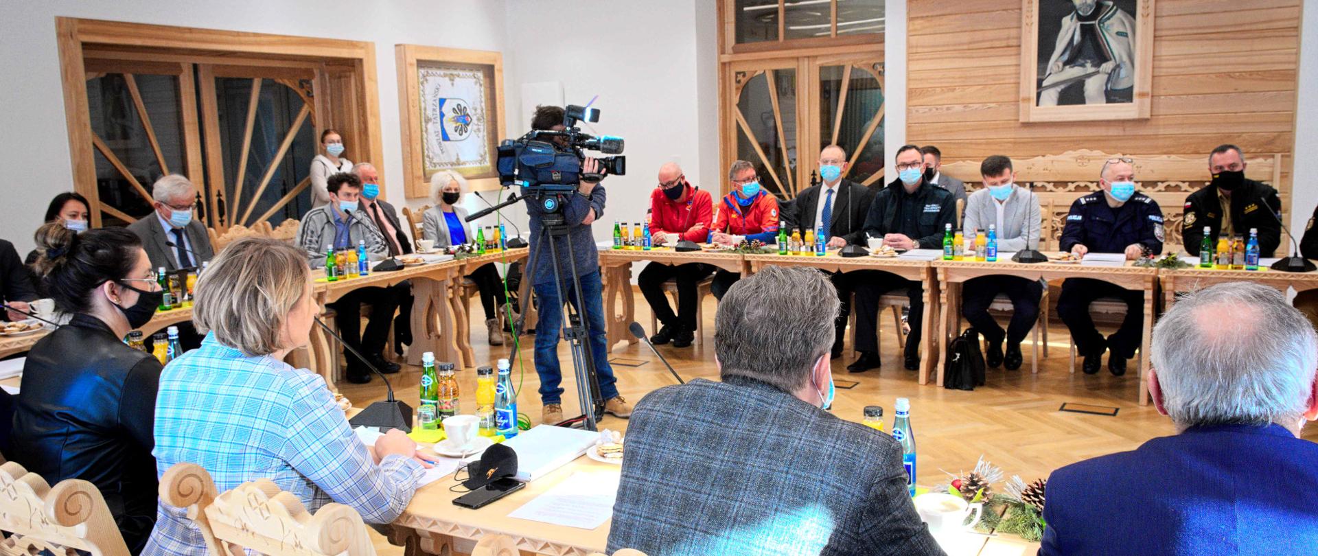Przedstawiciele służb siedzą dookoła stołu. Po środku operator z kamerą