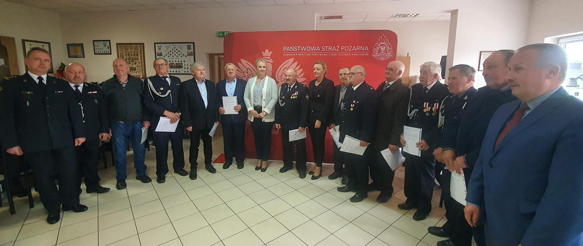 Wspólne zdjęcie druhów trzymających w rękach decyzję o przyznaniu świadczenia ratowniczego ze znamienitymi gośćmi biorącymi udział w uroczystości, Zdjęcie wykonane na tle baneru Państwowej Straży Pożarnej w Krotoszynie 