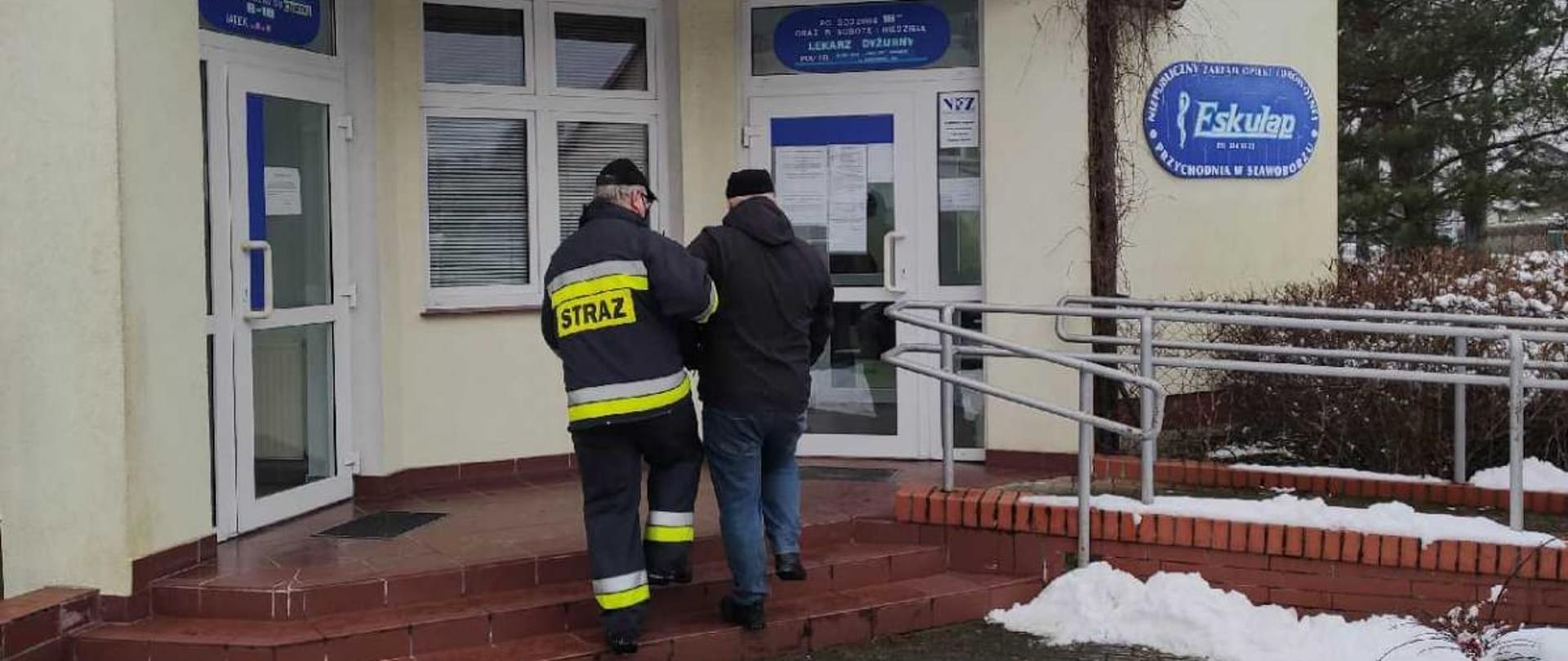 Zdjęcie przedstawia przychodnie zdrowia (punktem szczepień przeciwko COVID-19), otoczenie jest pokryte śniegiem. Strażak pomaga wejść starszemu mężczyźnie na schody, które prowadzą do wejścia przychodni. W tle zdjęcia widzimy drzewa oraz barierkę. 