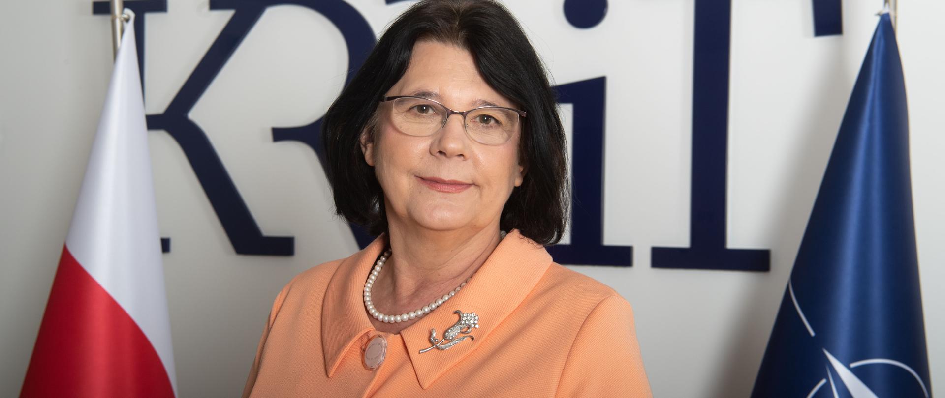 Portret Hanny Karp. Po lewej stronie flaga biało-czerwona, po prawej flaga NATO. Z tyłu niebieskie logo KRRiT.