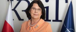 Portret Hanny Karp. Po lewej stronie flaga biało-czerwona, po prawej flaga NATO. Z tyłu niebieskie logo KRRiT.