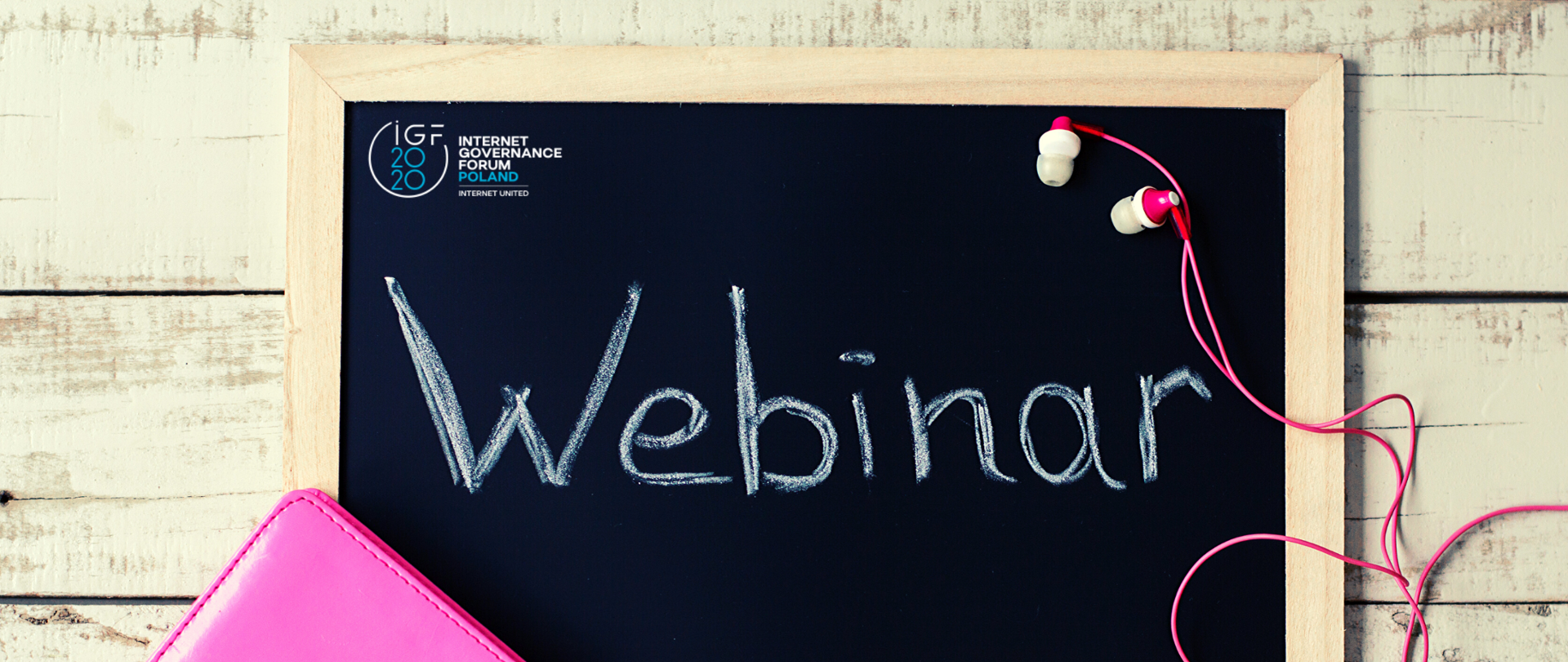 Na drewnianych deskach leży tablica przeznaczona do pisania kredą. Na tablicy napis: webinar, nad nim widoczne jest logo IGF 2020. W lewym dolnym rogu widać kawałek notesu w skórzanej różowej okładce. Po prawej stronie na tablicy leżą słuchawki z różowym kablem.