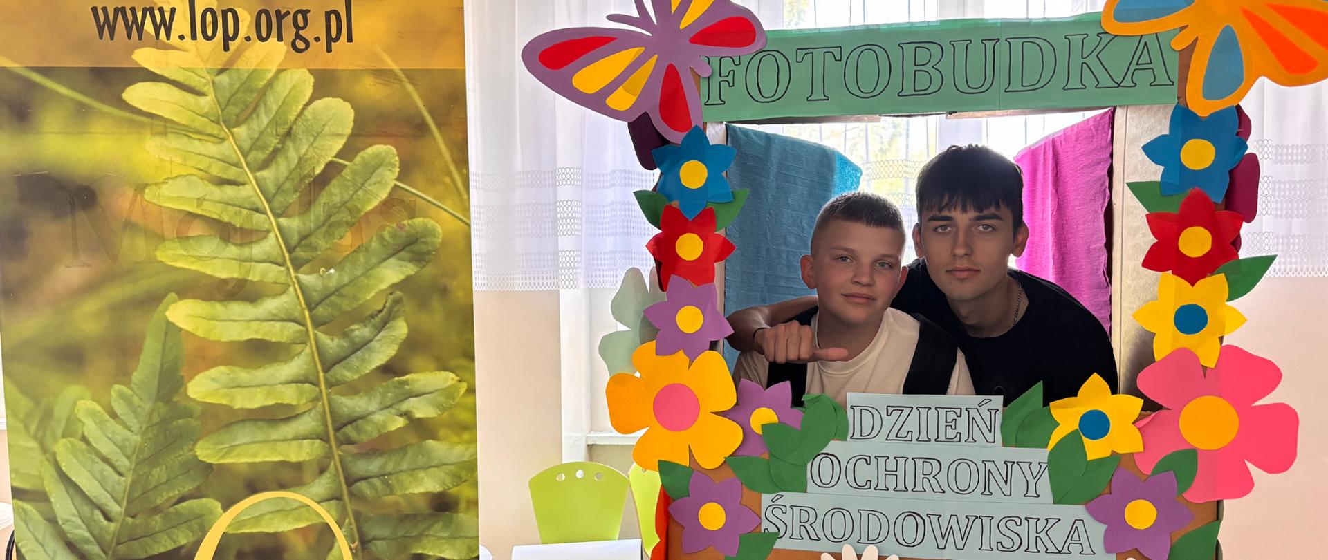 Dwie osoby pozują w fotobudce podczas wydarzenia o ochronie przyrody, obok widoczny baner informacyjny.