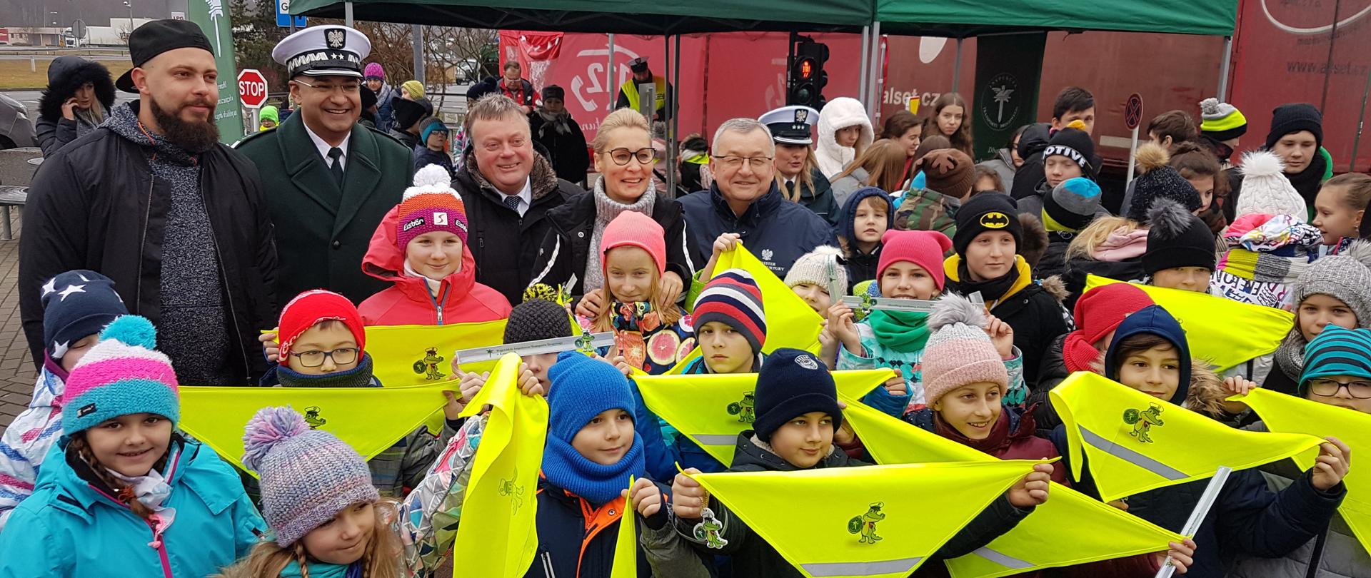 Na zdjęciu widać grupę dzieci, które trzymają chusty odblaskowe. W tle widać namioty reklamowe. 