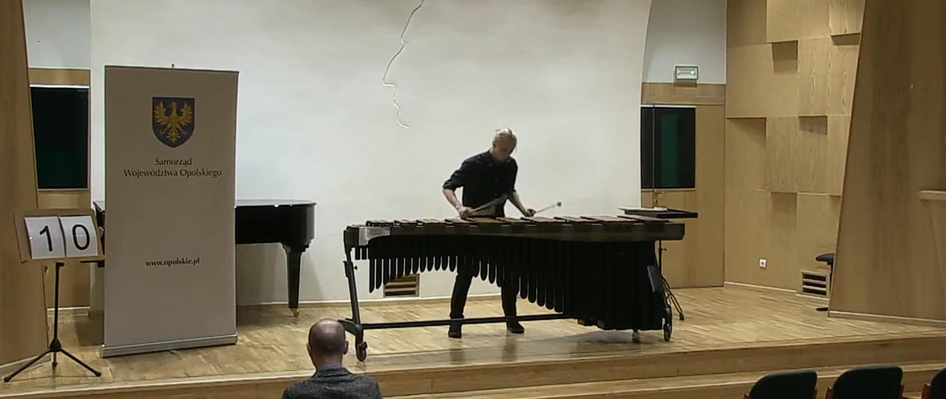 Sala kameralna szkoły muzycznej w Opolu scena na białym tle marimba i uczeń Filip Brzeziński grający w Czarnej koszuli obok baner reklamujący Urząd Województwa Opolskiego oraz pulpit z numerem 10