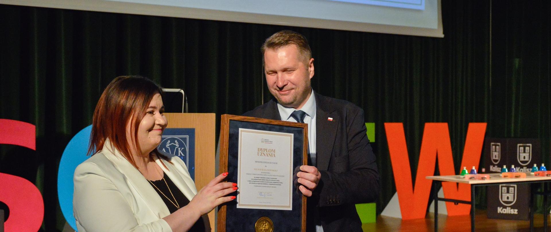 Minister Czarnek stoi obok kobiety w żółtej bluzce, razem trzymają oprawiony w ramki dokument, za nimi ściana zasłonięta ciemnozieloną kotarą.