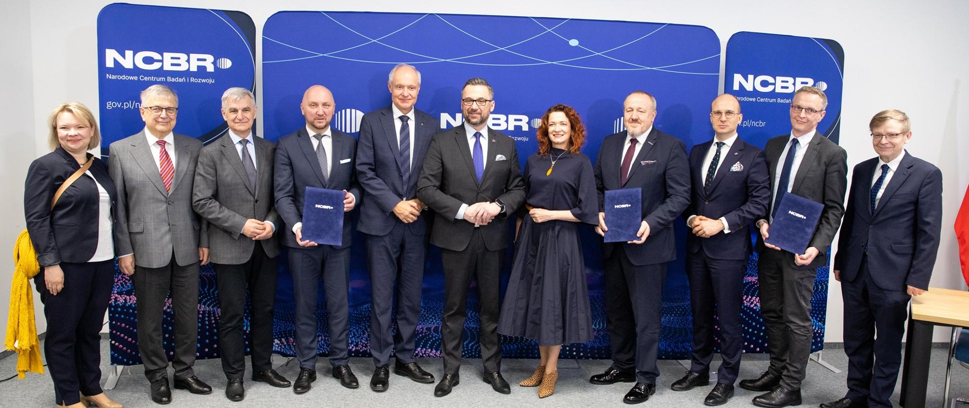 Zdjęcie zbiorowe - przed granatową ścianką z napisem NCBR stoi grupa jedenastu osób, pośrodku minister Kulasek.