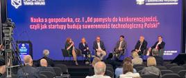 Zdjęcie od tyłu sali, na podwyższeniu z przodu na krzesłach siedzi sześć osób, kobieta z lewej mówi do trzymanego w ręku mikrofonu, za nimi na fioletowej ścianie napis Nauka a gospodarka cz. 1.