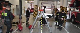 Na zdjęciu widzimy strażaków ubranych w stroje koszarowe koloru czarnego, na plecach napis straż. Strażacy mają także na sobie szelki wysokościowe. Akcja ma miejsce w garażu remizy, na środku kadru widzimy trójnóg. 