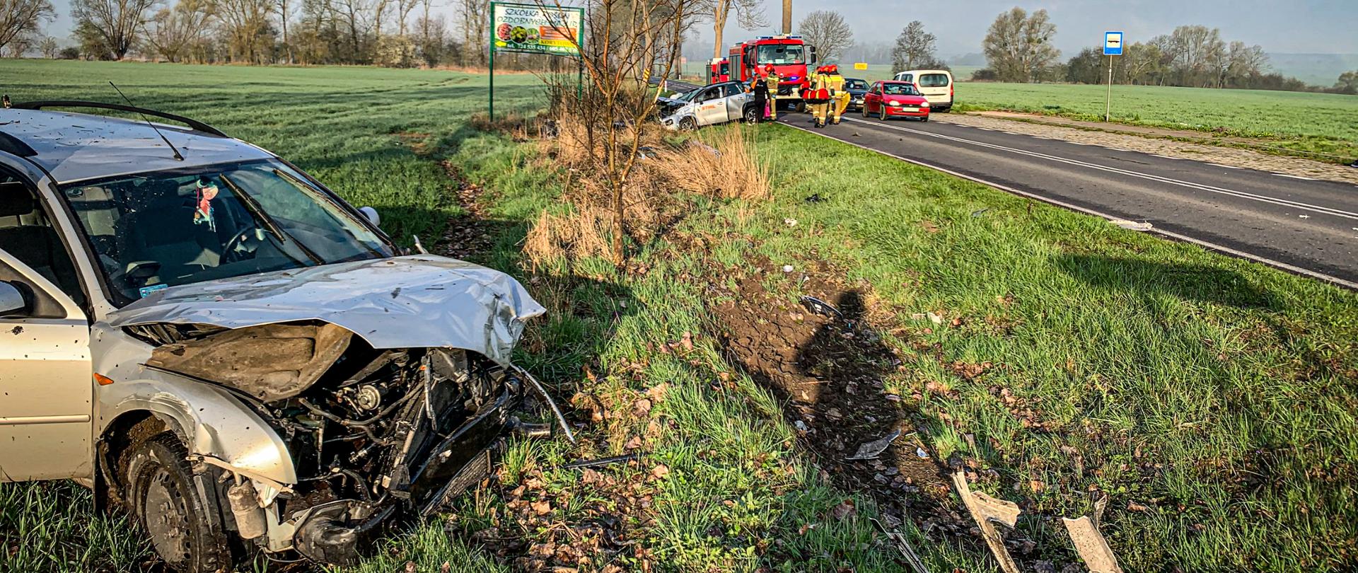 Na pierwszym planie zdjęcia po lewej stronie znajduję się rozbity przód samochodu osobowego po prawej stronie przebiega droga. Na drugim planie na poboczu drogi znajduje się drugi rozbity samochód uczestniczący w wypadku, wozy straży pożarnej jak i ratownicy. Zdjęcie zostało wykonanie w porze dziennej w słoneczny dzień. Na niebie przemieszczają się obłoki chmur.