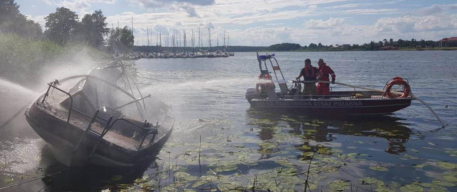 Dwaj strażacy chłodzący butle z gazem znajdujące się na spalonej łodzi.
