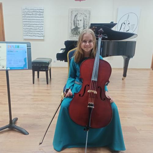 Na zdjęciu siedzi dziewczynka trzymając wiolonczelę obok ustawiono pulpit z Dyplomami w tle z napisami Dyplom Laureata i Dyplom za zajęcie II miejsca.. W tle fortepian i portrety Jana Sebastiana Bacha, oraz Logo Szkoły i partytura J. S. Bacha.