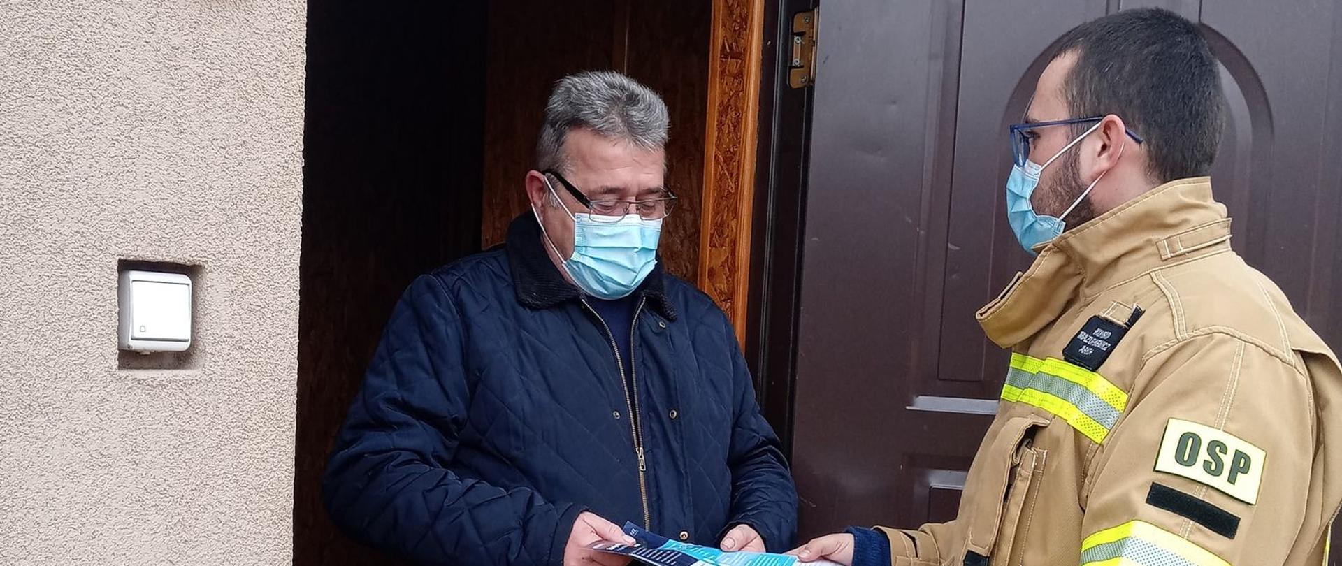Zdjęcie przedstawia strażaka w piaskowym ubraniu strażackim, który wręcza mężczyźnie ulotki #szczepimysię