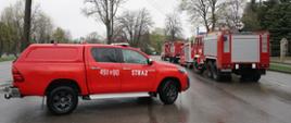 Zdjęcie przedstawia ciąg samochodów strażackich stojących jeden za drugim, które czekają na rozpoczęcie ćwiczeń. W tle widać parking kościoła parafialnego w Żołyni.