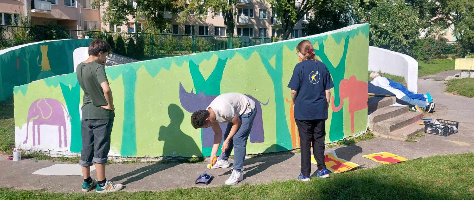 Jest ciepły, słoneczny dzień. Troje uczniów maluje murar (drzewa, słonie nosorożec) na murze przedszkola. W tle drzewa, ogrodzenie i bloki.