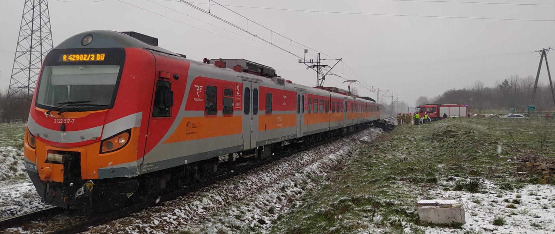 Po lewej stronie pociąg osobowy, po prawej stronie w tle samochód strażacki, strażacy PSP i Policjanci. na 1 planie śnieg na trawie. 