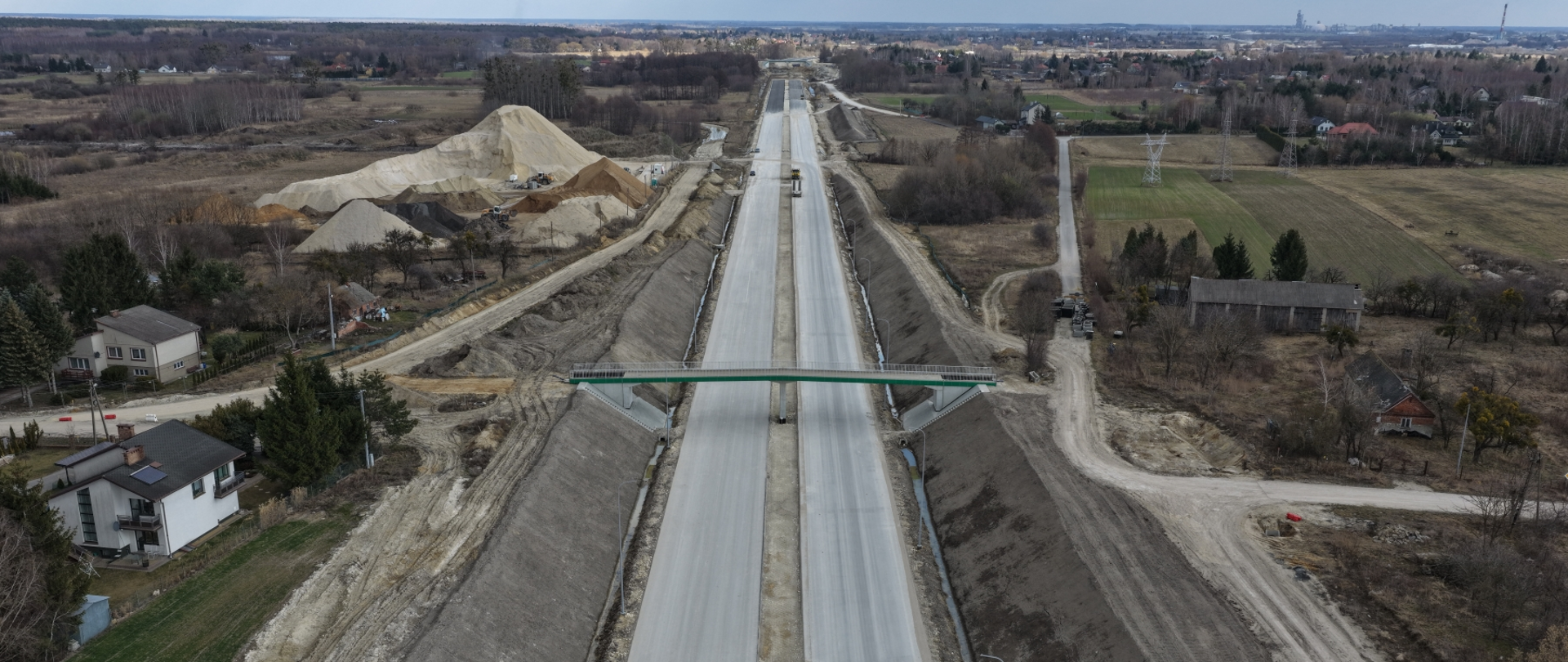 Widok z lotu ptaka na realizowany odcinek obwodnicy Chełma. Na środku kadru znajduje się dwujezdniowa trasa główna w budowie z wykonanymi nasypami oraz wiadukt drogowy KP‑010 w km 9+200. W otoczeniu widoczne są drogi dojazdowe, zabudowa jednorodzinna oraz tereny rolnicze.