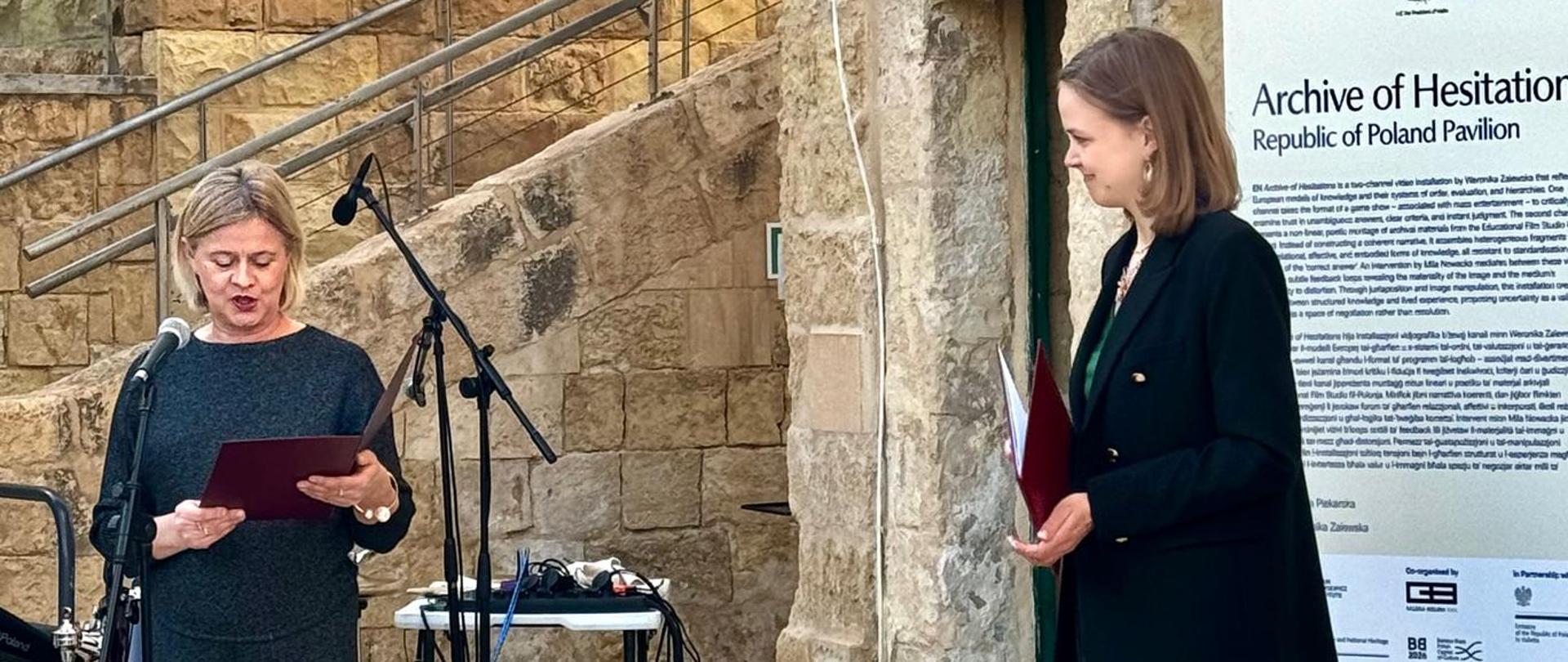 Pani Bożena Żelazowska, Sekretarz Stanu w Ministerstwie Kultury, czyta przemówienie podczas otwarcia Polskiego Pawilonu na Malta Biennale 2026. Przy drugim mikrofonie stoi profilowo druga kobieta, oczekująca na swoją kolej.