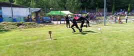 Zdjęcie przedstawia strażaków OSP biegnących podczas sztafety i podających sobie prądownice. Druhowie ubrani są w czarne stroje koszarowe, i biały hełm. Biegną po boisku z trawy.