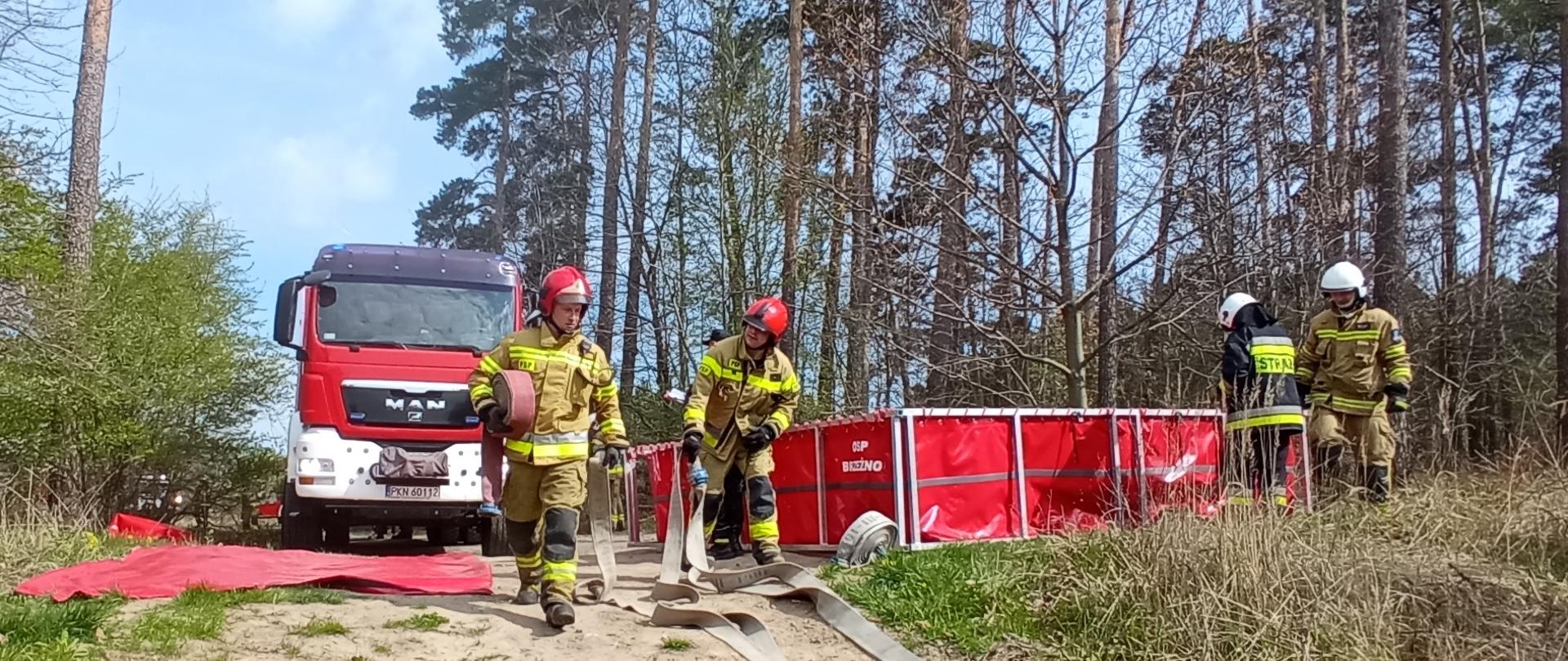 Na pierwszym planie widać dwóch strażaków z Państwowej Straży Pożarnej rozwijających węże pożarnicze. Po prawej stronie dwóch druhów z OSP rozkłada zbiornik brezentowy na wodę. W tle widać samochód pożarniczy