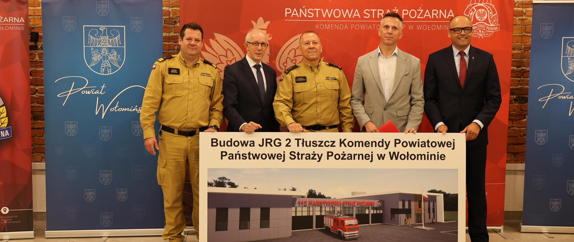 Na zdjęciu uczestnicy uroczystości prezentują planszę z wizualizacją przyszłej Jednostki Ratowniczo-Gaśniczej w Tłuszczu. Po bokach stoją strażacy PSP w piaskowych mundurach służbowych oraz przedstawiciele administracji publicznej w garniturach. W tle znajdują się banery Komendy Powiatowej PSP w Wołominie i Powiatu Wołomińskiego.