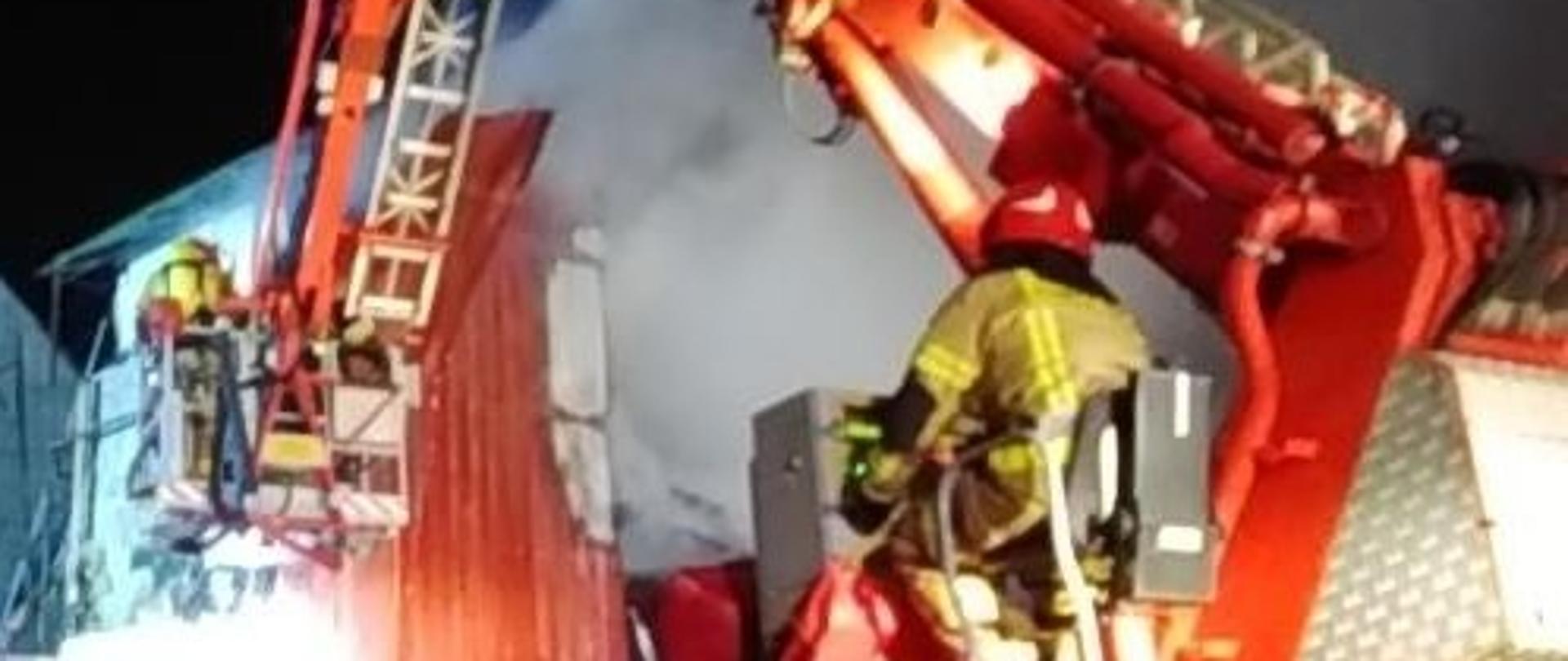 Funkcjonariusz operujący podnośnikiem, w tle pożar suszarni zboża oraz funkcjonariusze w koszu podnośnika