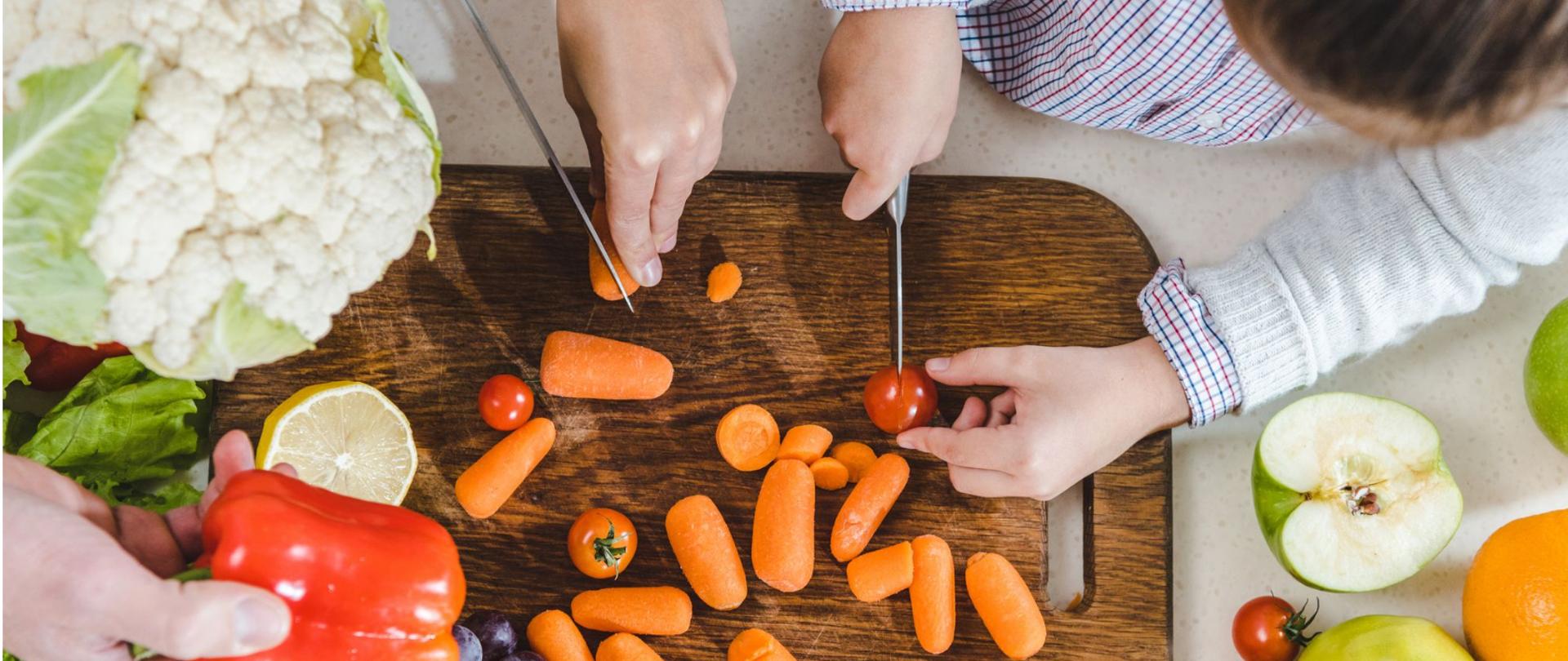 Na zdjęciu widoczne są dwie osoby, które kroją warzywa na drewnianej desce. Trzecia osoba trzyma w dłoni paprykę.