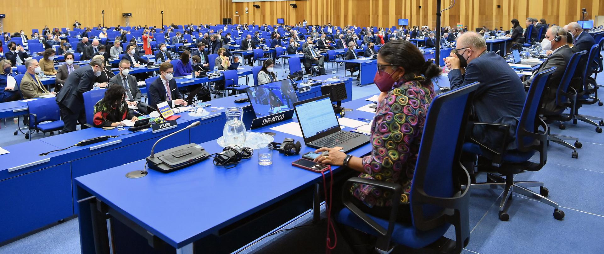 Nadzwyczajne posiedzenie Rady Gubernatorów MAEA - sala plenarna. Uczestnicy siedzą przy niebieskich stołach. Na pierwszym planie kobieta ubrana w kolorowy żakiet patrzy się w ekran laptopa.