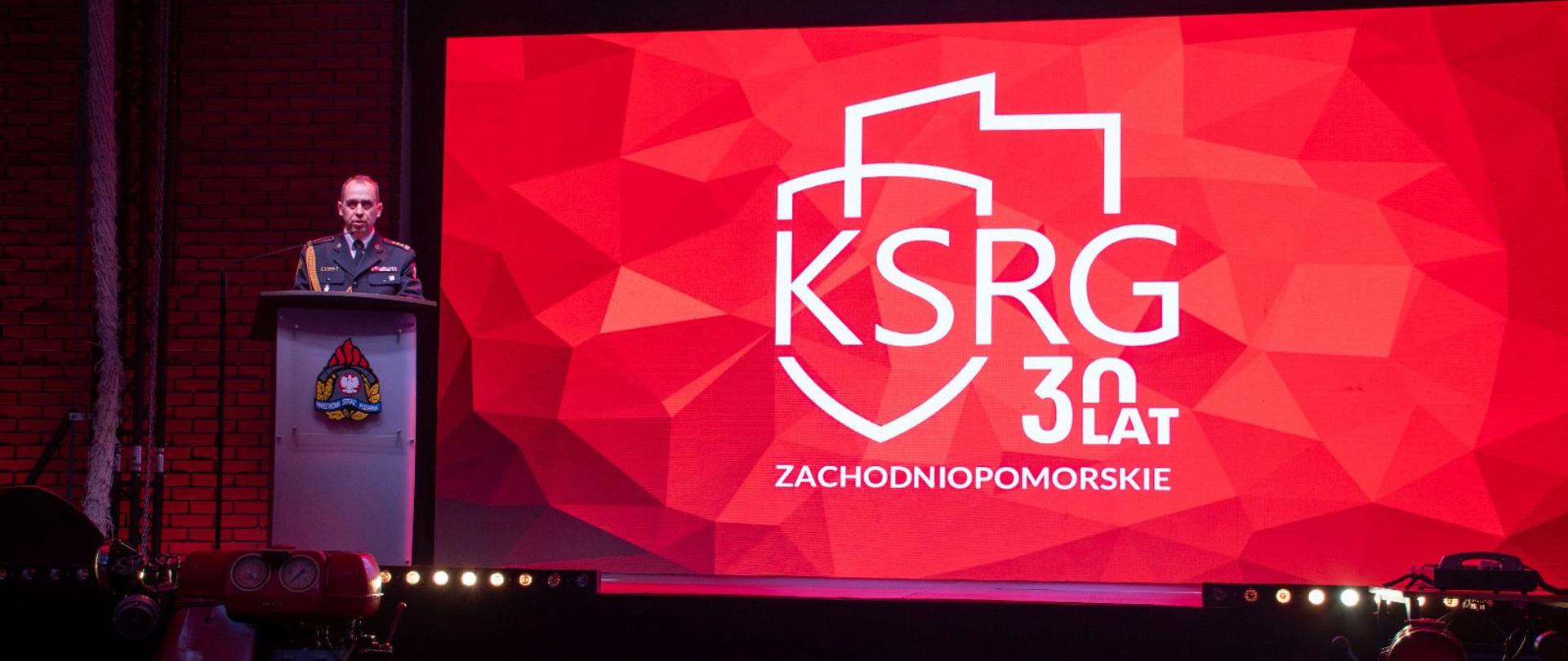 Na zdjęciu widać scenę wewnątrz dużej, ciemnej sali. Przestrzeń oświetlona jest głównie czerwonym światłem. Po lewej stronie sceny stoi mównica. Za nią znajduje się umundurowany strażak w galowym stroju, widoczny od pasa w górę. Na frontowej części mównicy umieszczony jest kolorowy herb Państwowej Straży Pożarnej.
Centralnym i największym elementem kadru jest szeroki ekran LED o intensywnie czerwonym, geometrycznym tle. Na ekranie widnieje biały logotyp składający się z konturu stylizowanej tarczy oraz dużego napisu „KSRG”. Obok niego znajduje się biały napis „30 LAT”. Pod logotypem widać mniejszy napis „ZACHODNIOPOMORSKIE”.
Przed sceną, po lewej i prawej stronie, stoją elementy sprzętu strażackiego, między innymi motopompy i zwinięte węże. Cała scena sprawia wrażenie oficjalnego wydarzenia z okazji rocznicy funkcjonowania systemu ratowniczo–gaśniczego.