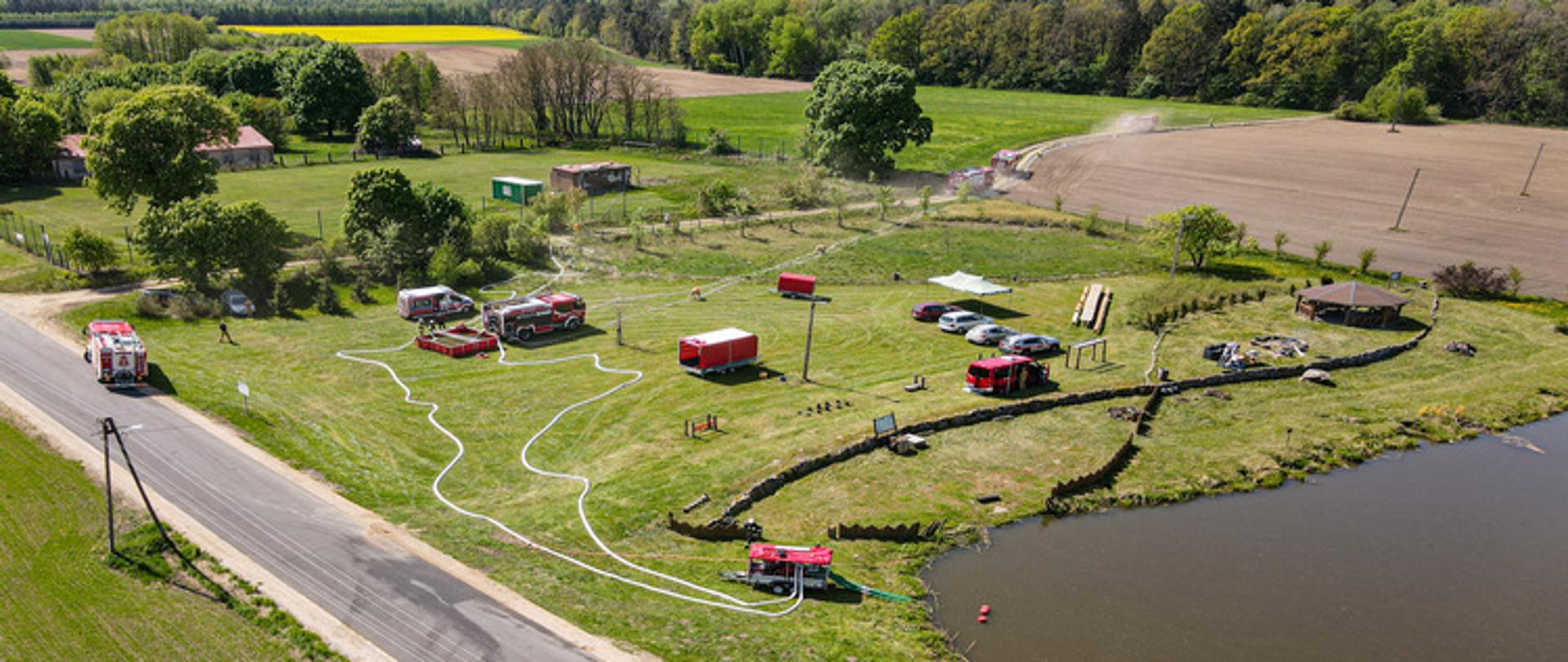 To zdjęcie to ujęcie z lotu ptaka (dronu) przedstawiające obszerny, zielony teren łąkę, zaadaptowaną na potrzeby ćwiczeń. Widoczna jest duża ilość sprzętu gaśniczego i ćwiczebnego.
Na pierwszym planie, po prawej stronie, znajduje się staw lub zbiornik wodny. Z tego zbiornika biegną białe węże strażackie, podłączone do pomp, rozłożone po trawiastym terenie. Na polu rozmieszczone są różne elementy treningowe, w tym czerwone baseny lub zbiorniki na wodę, mniejsze czerwone naczepy lub przyczepy, a także kilka wozów strażackich. W centrum kadru widać także zaparkowane samochody osobowe.
Po lewej stronie biegnie droga asfaltowa, a za nią widoczne są wiejskie zabudowania. W oddali, w prawym górnym rogu, widać pola uprawne, w tym jedno z żółtą uprawą (prawdopodobnie rzepakiem). Scena jest otoczona zielonymi drzewami i krzewami, a nad całością rozpościera się jasne niebo. Całość to rozległe ćwiczenia, gdzie testowana jest sprawność i koordynacja działań strażaków w terenie.