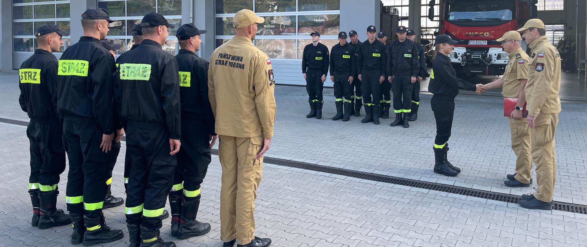 Na zdjęciu Komendant Powiatowy PSP w Śremie wręcza strażakowi list gratulacyjny obok stoi Zastępca Dowódcy Jednostki a na przeciwko siebie zmiany służbowe strażacy w ubraniach koszarowych za nimi otwarta brama garażowa w której stoi samochód ratowniczo - gaśniczy