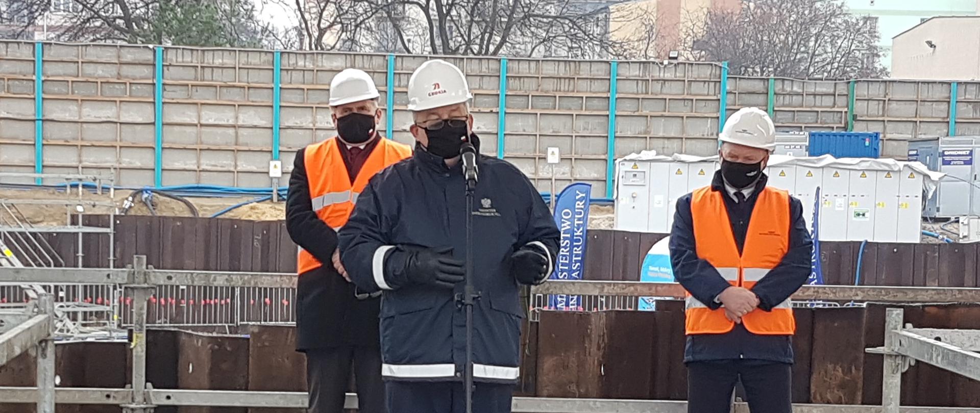 Na zdjęciu widać 3 osoby, które są na placu budowy. Są to mężczyźni, którzy ubrani są w jasne kamizelki, na głowach mają kaski.