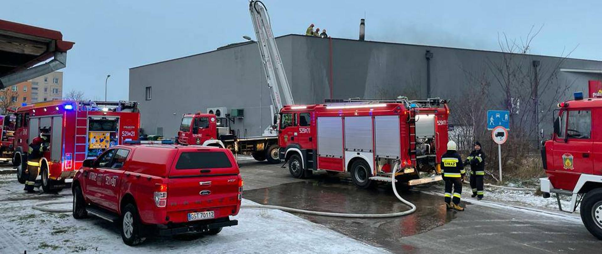 Zdjęcie przedstawia miejsce prowadzonych działań, na pierwszym planie samochody pożarnicze, samochody na sygnałach świetlnych, strażacy na dachu budynku